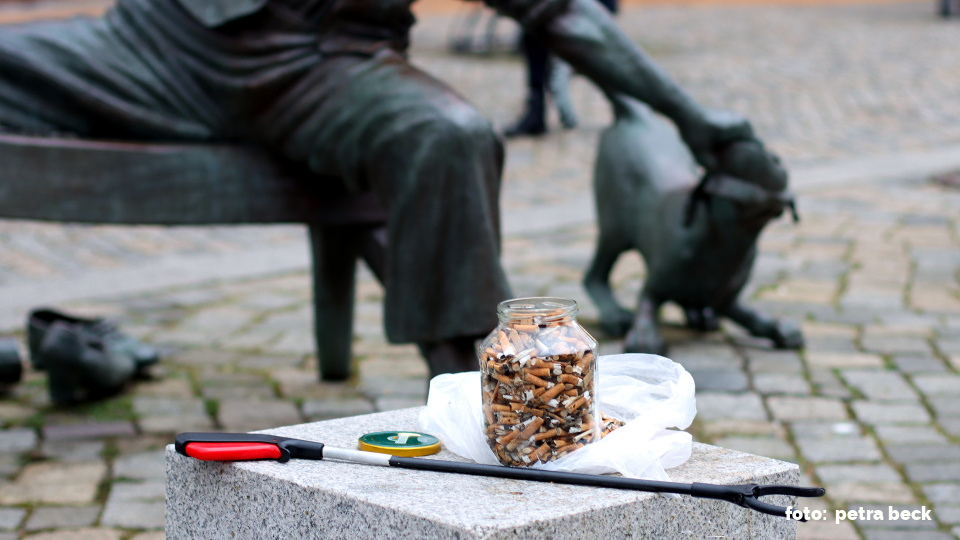 Vor einer Statue eines sitzenden Schusters, der mit einem kleinen Dackel (auch als Statue) um einen Schuh kämpft, steht ein Glas mit aufgesammelten Zigarettenkippen. Diese hatte ich auf dem Marktplatz in Preetz gesammelt.