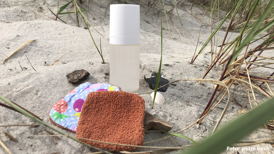 Im Sand am Ostseestrand liegen zwei selbst genähte Abschminkpads aus alten Handtüchern und buntem Baumwollstoff vor einer Flasche mit einem Kosmetikprodukt. rechts davon ist Strandhafer zu sehen, der aus dem Sanf wächst.