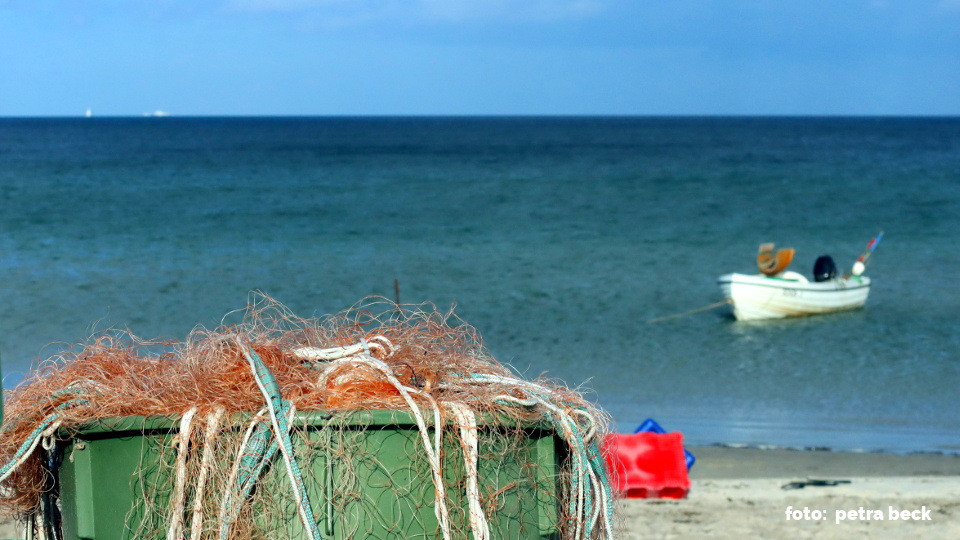 Am Strand steht eine Tonne mit Fischernetzen aus Plastik. Im Hintergrund schwimmt ein kleines Fischerboot im Meer.