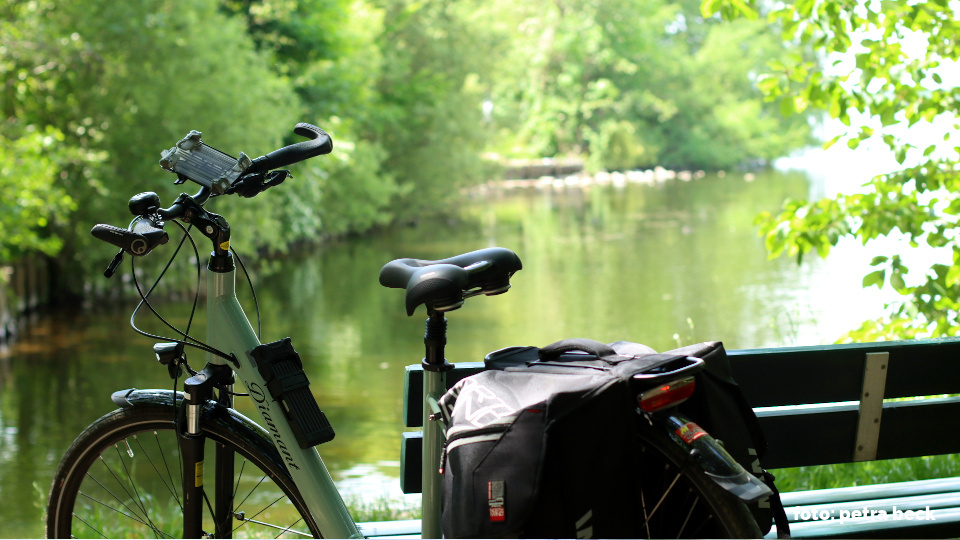 En Fahrrad steht neben einer Bank. Im Hintergrund befindet sich ein See der mit Büschen gesäumt ist. Es entstand während der Pause einer Fahrradtour. 